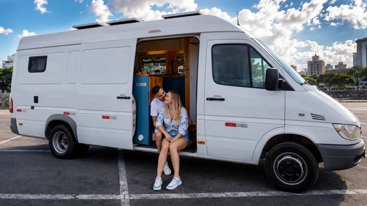 Casal sentado na porta de um motorhome branco estacionado no Estacionamento Salvador, no centro de Salvador-BA, com interior moderno visível e ambiente seguro para viajantes.